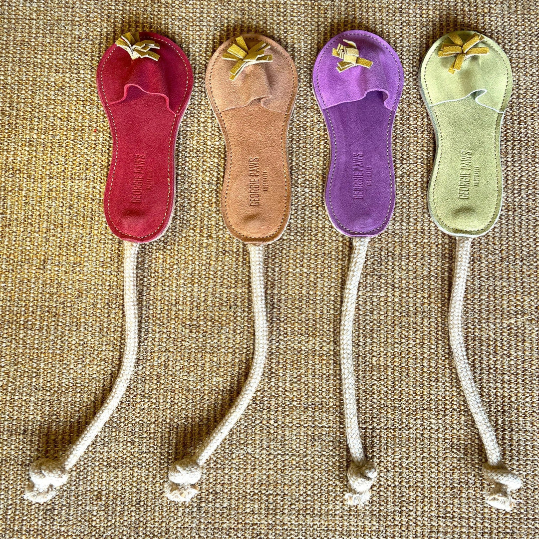 Four vibrant Sigrid Slipper bookmarks from Georgie Paws—in red, tan, purple, and green—each handmade from vegetable-tanned buffalo leather with a knotted rope strap and decorative tassel, are arranged neatly on a tan woven surface.