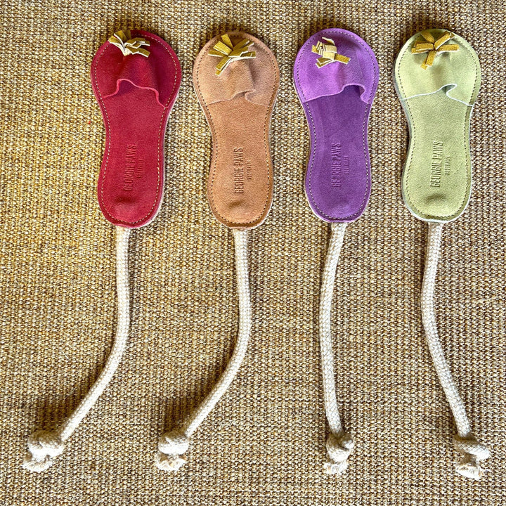 Four vibrant Sigrid Slipper bookmarks from Georgie Paws—in red, tan, purple, and green—each handmade from vegetable-tanned buffalo leather with a knotted rope strap and decorative tassel, are arranged neatly on a tan woven surface.