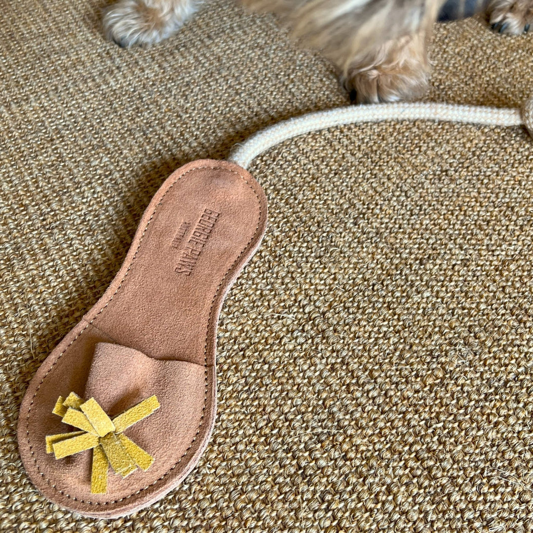 A Sigrid Slipper - Natural dog toy by Georgie Paws, crafted from natural vegetable tanned buffalo leather and shaped like a brown slipper with a yellow bow and the word "Souvenir," rests on a tan woven rug. A rope is attached to the toy, and part of a small dog's paws and fur appear at the top of the image.
