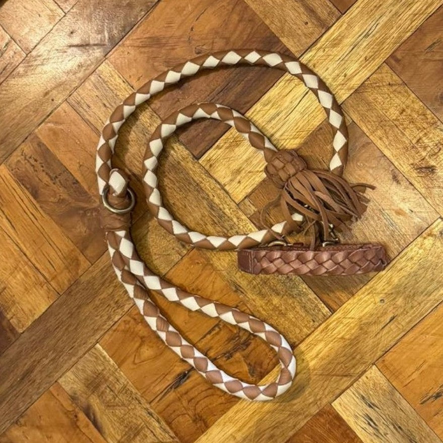 The Georgie Paws Casey Collar - Syrup, a braided tan and cream leather dog leash with brass hardware, loop handle, metal clasp, and decorative tassel, is coiled on a herringbone parquet wooden floor.