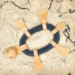 A Thomas the Turtle - natural & pink child's toy made of buffalo suede and fabric rests on sandy ground, suggesting playful outdoor activities possibly at a beach or sandbox. This product is from Georgie Paws.