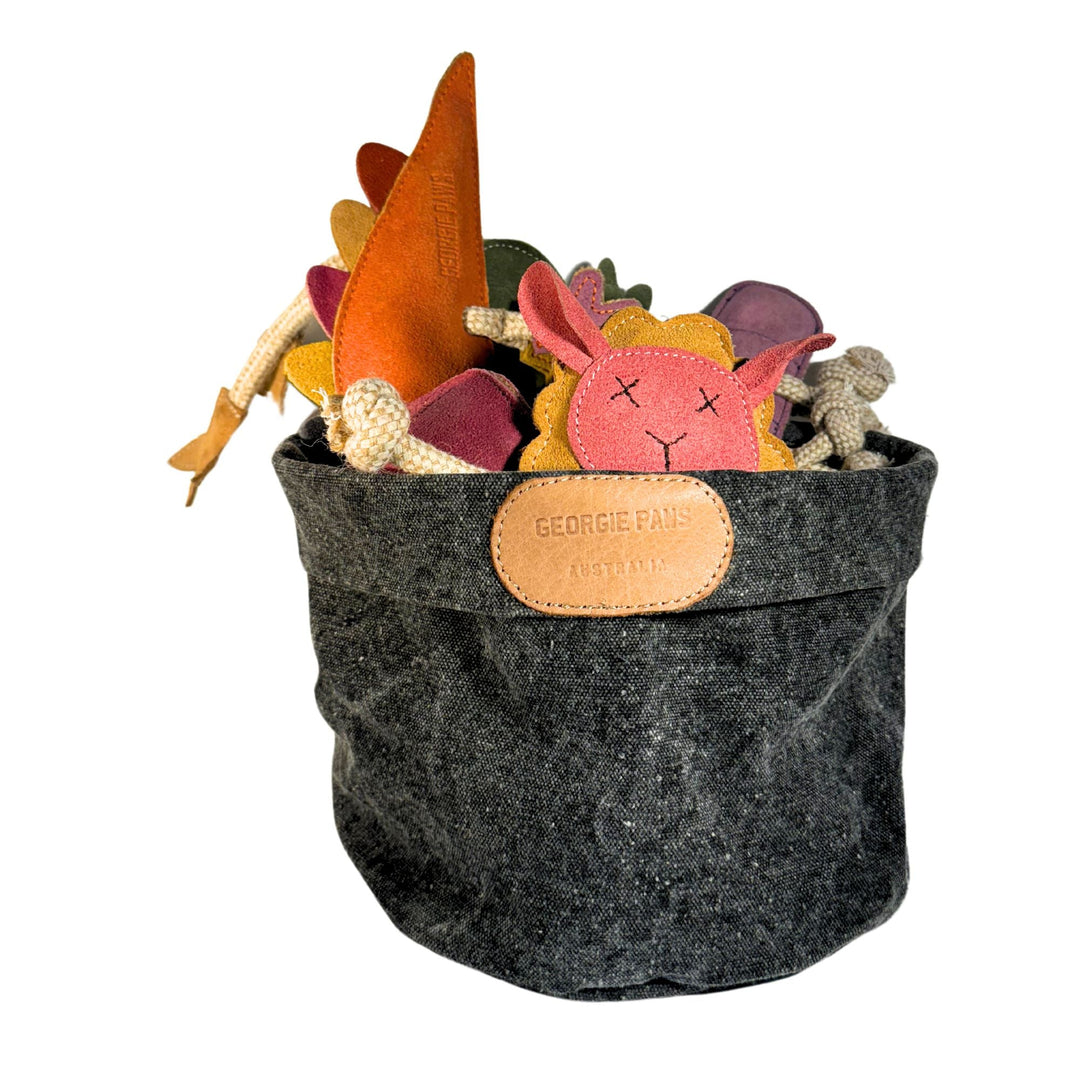 The Georgie Paws Toy Storage Bucket is a dark gray fabric basket adorned with a tan vegetable-tanned buffalo leather patch that reads "GEORGIE PAWS AUSTRALIA." It contains an assortment of colorful felt animal toys, such as a pink lion face with a yellow mane. Made from sustainable materials, this storage bucket is pictured against a white background.