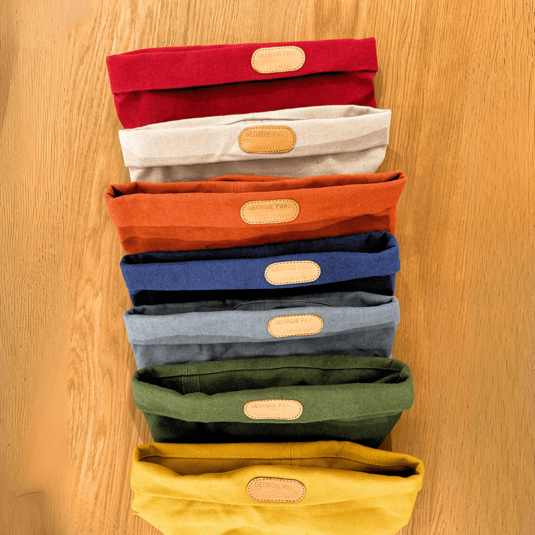 Eight folded Georgie Paws Toy Storage Buckets in a range of colors (red, beige, orange, rust, blue, light gray, green, and yellow) are stacked vertically on a wooden surface. Each sustainable toy storage bucket features a tan oval patch with faint text centered near the top fold.