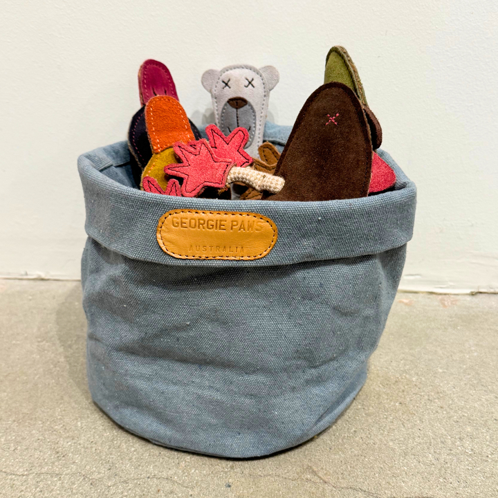 The Georgie Paws Toy Storage Bucket holds colorful felt dog toys—white bear, brown bone, and pink shapes. This soft blue canvas basket features a branded label and is displayed on a concrete floor against a plain wall.