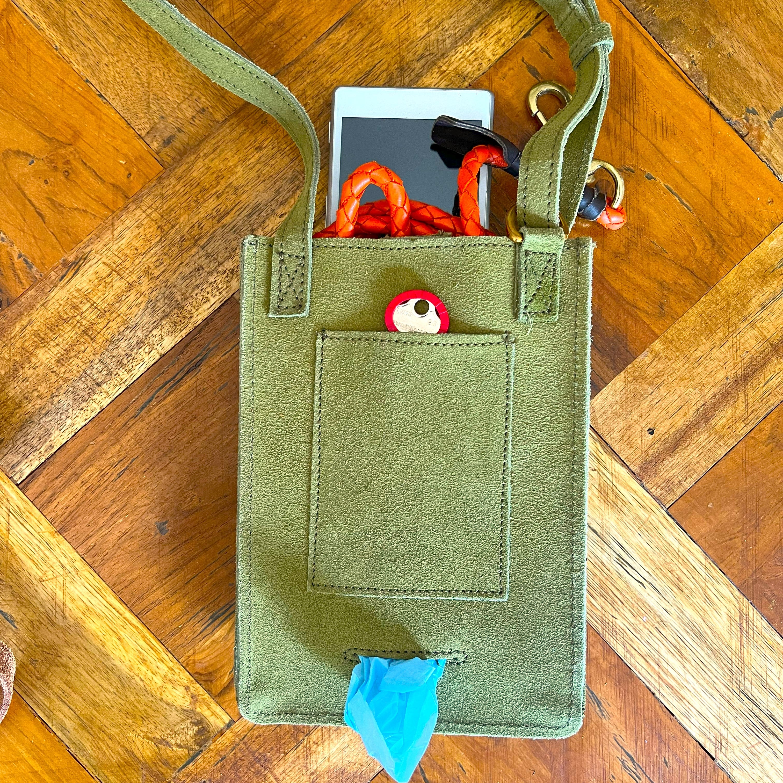 A grass green Dog Walk Bag crafted from natural veg tanned leather lying on a wooden floor, open to show items inside: a white smartphone, an orange scarf, a red and white spotted keychain, and