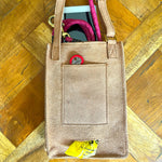 A tan Veg Tanned Leather tote bag with pink woven handles sits on a wooden floor. The Georgie Paws Dog Walk Bag - Raw is partially filled, showing a bright pink scarf, a red circular tag, and yellow packaging peeking