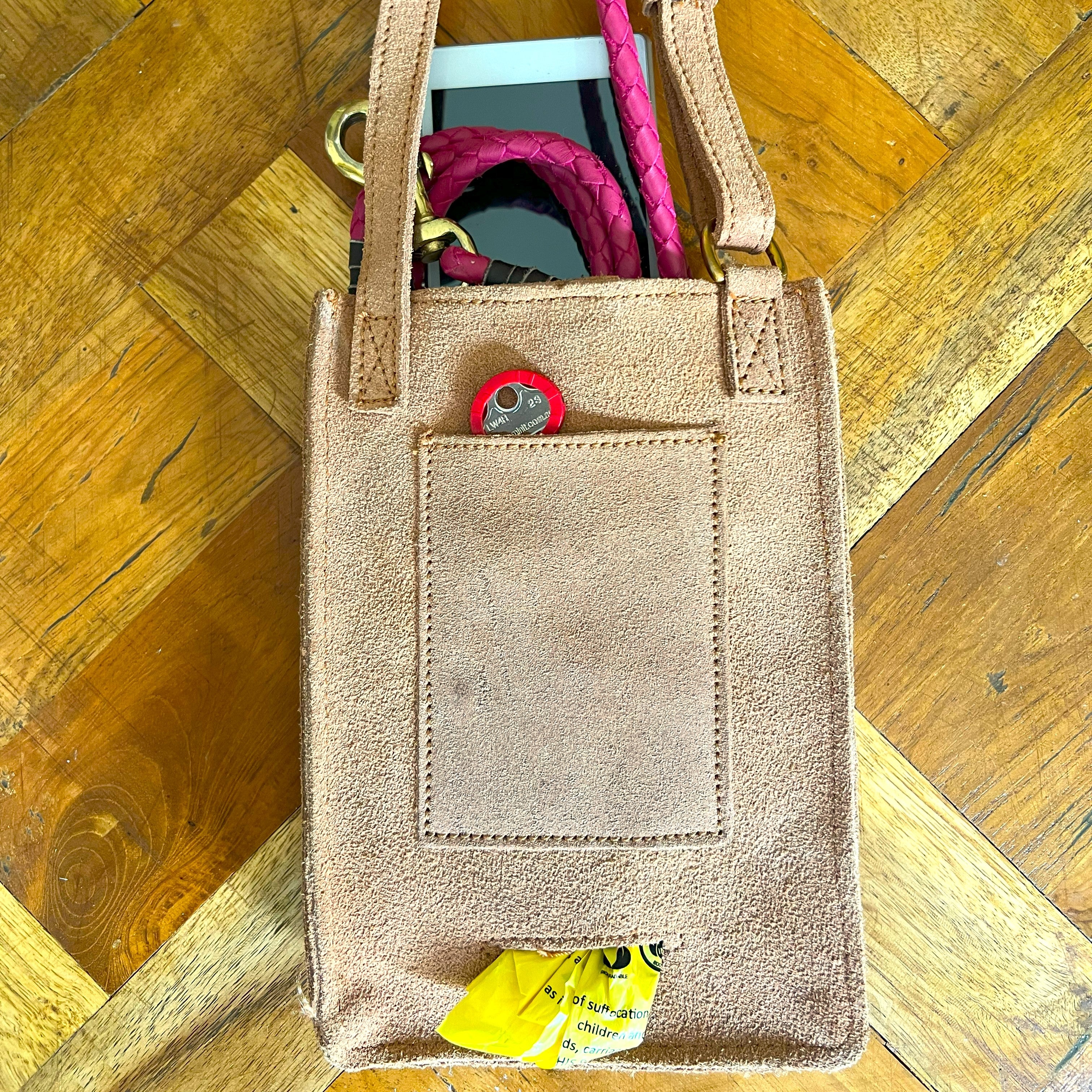 A tan Veg Tanned Leather tote bag with pink woven handles sits on a wooden floor. The Georgie Paws Dog Walk Bag - Raw is partially filled, showing a bright pink scarf, a red circular tag, and yellow packaging peeking
