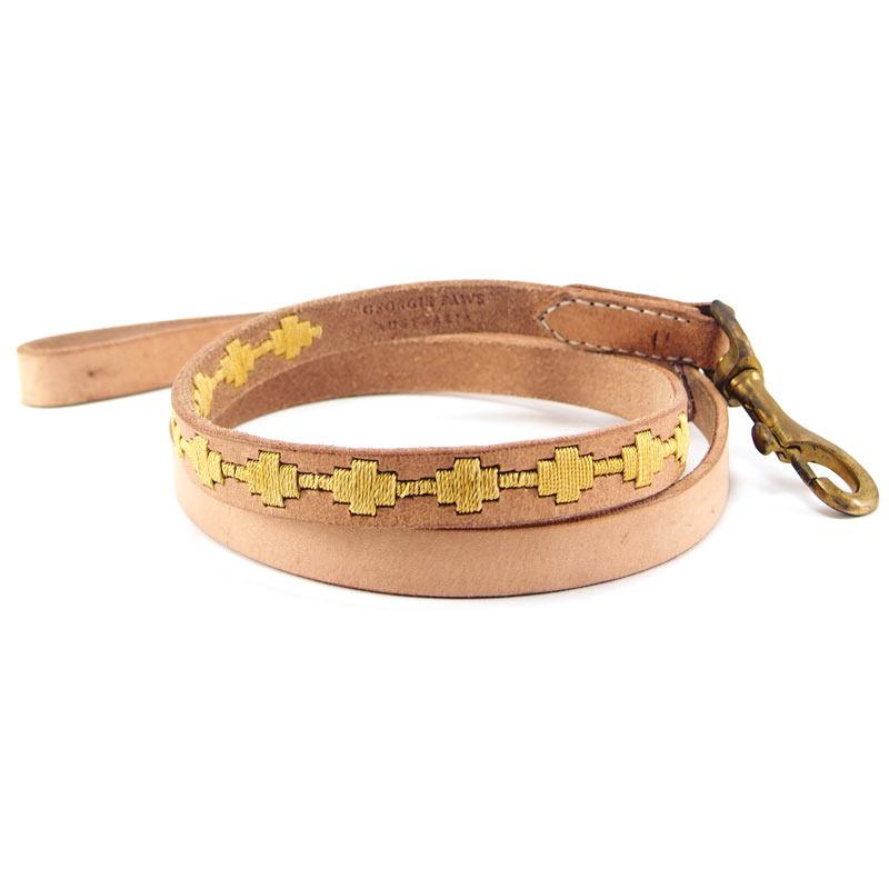 The Ethel Lead - Gold by Georgie Paws is a light tan, vegetable-tanned buffalo leather dog leash, coiled neatly and adorned with a pattern of yellow geometric shapes along its length. It features a brass swivel snap hook on one end and a simple loop handle on the other.