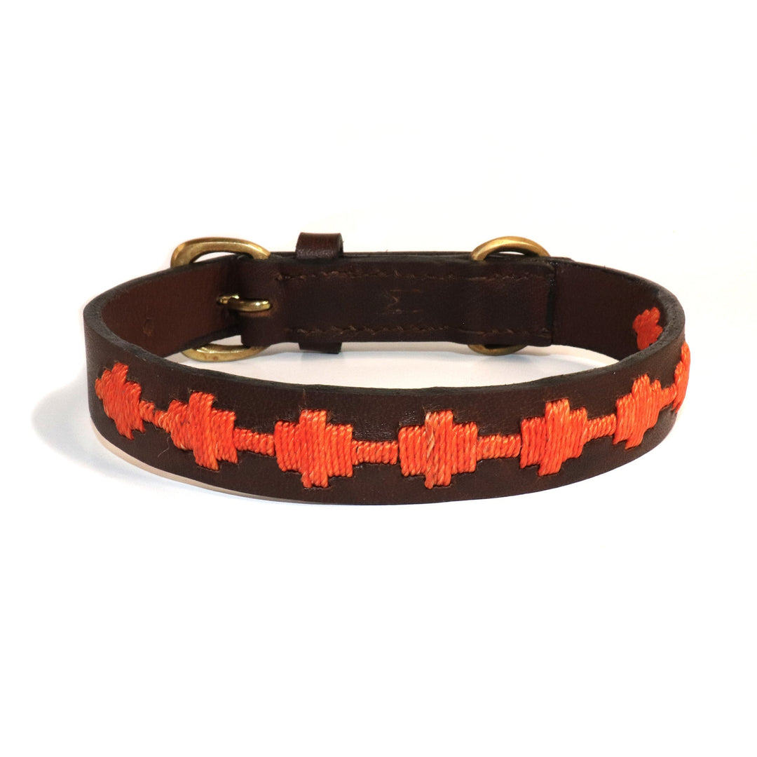 The Polo Bark Collar - Ochre by Georgie Paws is crafted from sustainable, vegetable-tanned buffalo leather in a rich brown shade with brass hardware. It features a striking pattern of orange geometric crosses embroidered along its length and is shown coiled in a circular shape on a white background.